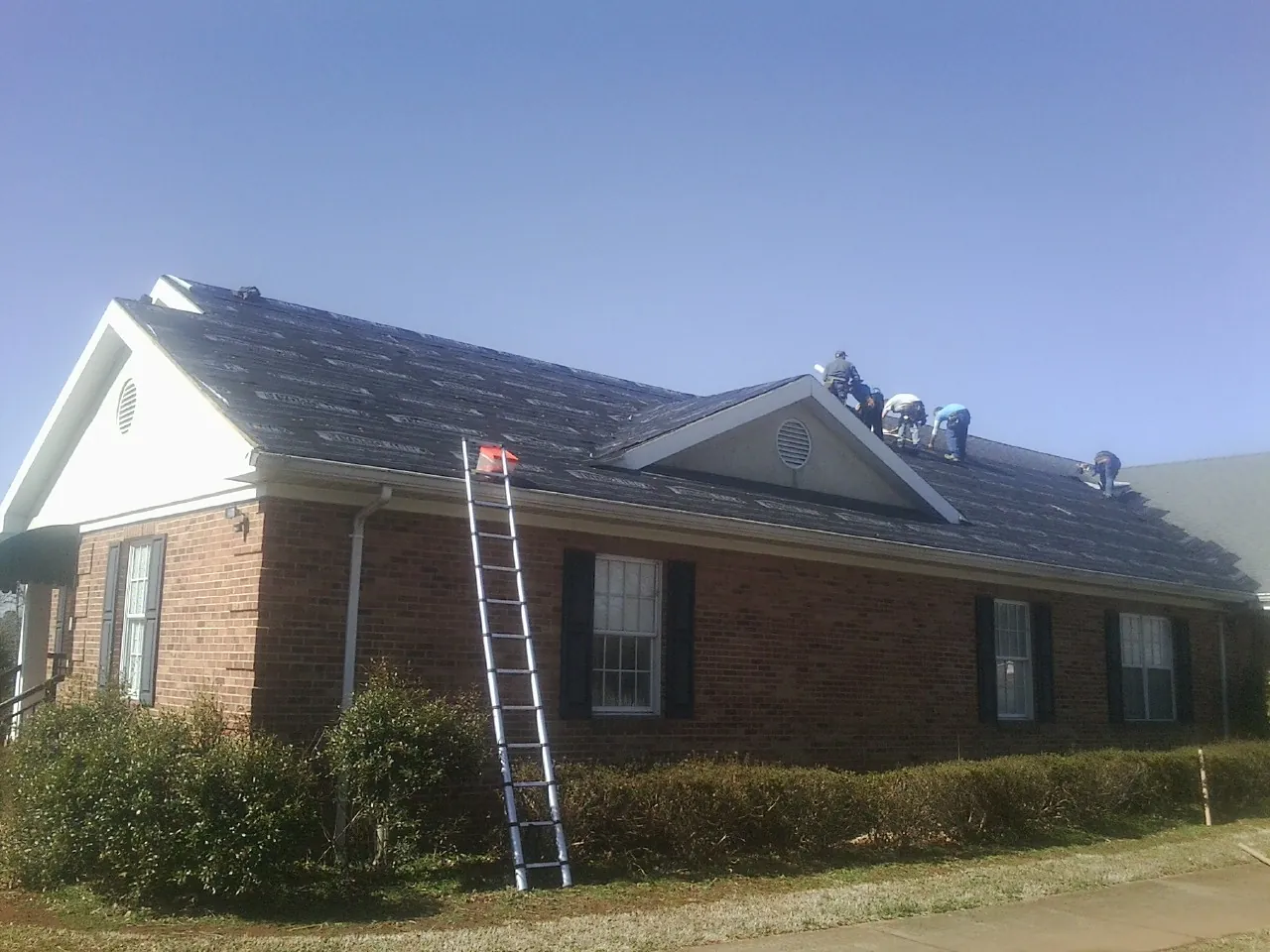 Crew tearing shingles off a brick ranch roof with a ladder set — Emergency Roof Repair work by our crew in Mathews