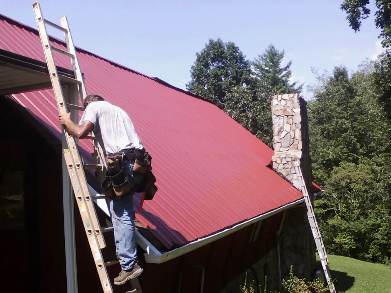 Roofer on a ladder beside a red metal roof and stone chimney — Soffit & Fascia Repair by Roofing Brothers in Mathews